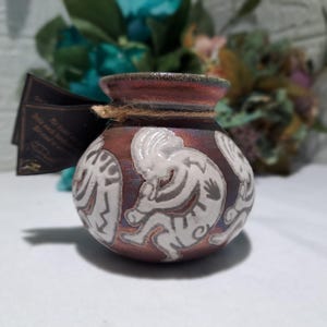 May include: A small, round, metallic-toned ceramic pot with a dark, iridescent finish. The exterior features white, embossed figures of flute players. A brown twine is tied around the neck, holding a dark rectangular tag with printed text.
