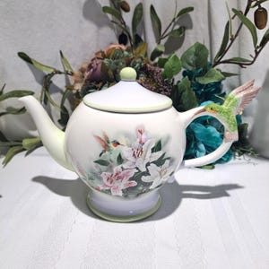 May include: A white ceramic teapot with a lid and a hummingbird-shaped handle. The teapot is decorated with a floral design featuring pink and white lilies and hummingbirds. The spout and base have a pale green trim.