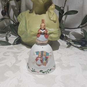 May include: A white ceramic bell with a green and red trim. The bell features a depiction of two rabbits under an umbrella. One rabbit is holding a bouquet of flowers and the other is holding a book.