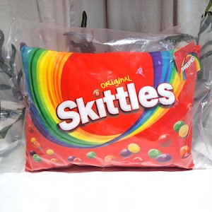 May include: A rectangular pillow featuring the Skittles candy logo. The pillow has a red background with a rainbow arc design and the word "Skittles" in large white letters. The pillow is packaged in clear plastic.
