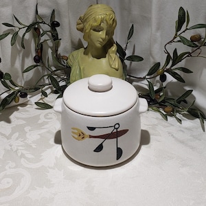 May include: White ceramic crock pot with a lid and a brown and yellow design of kitchen utensils on the front.