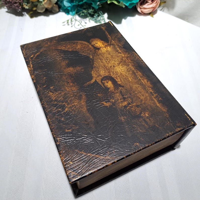 May include: A vintage-style book with a dark brown cover featuring a faded illustration of an angel and a person. The cover has a textured, distressed appearance. The book's edges are visible, suggesting aged paper.