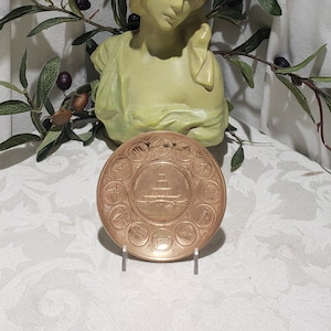 May include: A bronze-colored decorative plate with a central image of a building, surrounded by smaller circular images. The plate is displayed on a clear stand, with a statue and olive branch in the background.