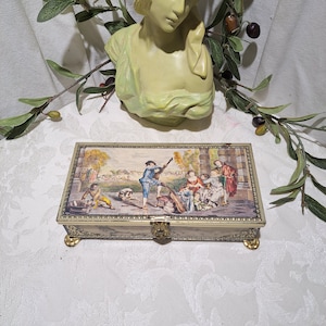 May include: A vintage metal trinket box with a hinged lid and gold accents. The box features a colorful illustration of people in a garden setting. The lid is closed.