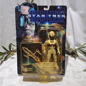 May include: A Star Trek: First Contact action figure of Captain Picard in his Starfleet spacesuit. The figure is holding a phaser rifle and is standing on a clear plastic base. The packaging is blue and yellow and features the Star Trek logo.
