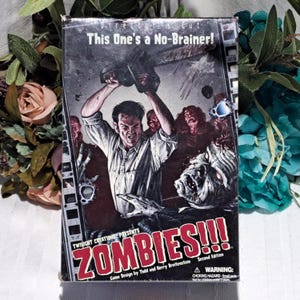 May include: A board game box titled "ZOMBIES!!!" features a man wielding a chainsaw against zombies. The box reads "Director's Cut, This One's a No-Brainer!" and "Second Edition." The game is designed by Todd and Kerry Breitenstein. A warning about choking hazards is also present.