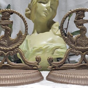 May include: A pair of antique brass bookends featuring a detailed anchor design. The anchors are surrounded by a rope-like border and sit on a brown base.