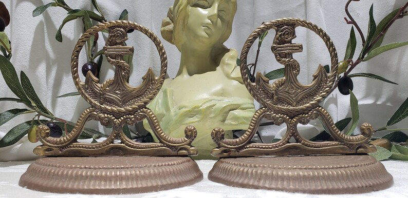 May include: A pair of antique brass bookends featuring a detailed anchor design. The anchors are surrounded by a rope-like border and sit on a brown base.