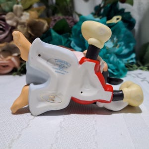 Pode incluir: Uma estatueta de porcelana de um personagem de desenho animado, com uma roupa vermelha e branca, detalhes em preto e grandes luvas amarelas. A estatueta est&aacute; deitada de costas, com texto vis&iacute;vel na base que diz "Disney & Me".