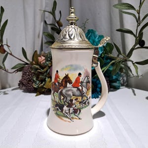 May include: Cream-colored ceramic beer stein with a silver-colored lid and handle. The stein features a hunting scene with riders on horseback and dogs. The lid has a decorative floral design.