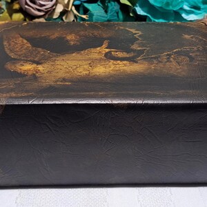 May include: A rectangular, dark brown box with a textured surface. The top features a faded, sepia-toned painting of a figure in a landscape. The box is likely for storage or display, with a vintage or antique aesthetic.