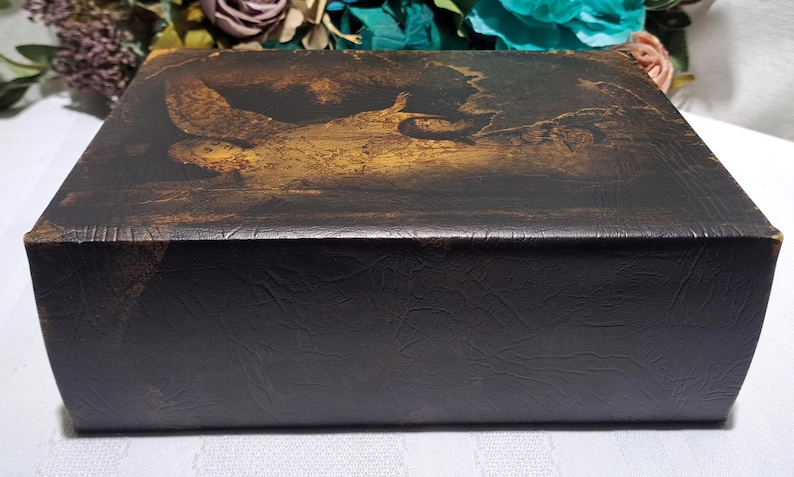 May include: A rectangular, dark brown box with a textured surface. The top features a faded, sepia-toned painting of a figure in a landscape. The box is likely for storage or display, with a vintage or antique aesthetic.