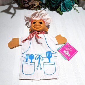 May include: A vintage troll doll wearing a chef's hat, a red and white checkered scarf, and a white apron with blue utensil designs. The doll has orange hands and a tag that says "Just say it with RUSS".