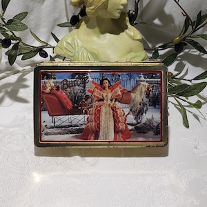 May include: A gold-colored metal tin with a Christmas scene featuring a Barbie doll in a red and gold gown riding in a red sleigh pulled by a white horse. The tin has a gold trim and the word "Barbie" is printed on the front.