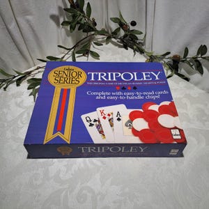May include: A blue box for the Cadaco Senior Series Tripoley game. The box features a red, white, and gold ribbon with the words "Cadaco Senior Series" in a gold circle. The box also features a picture of playing cards and chips. The text on the box reads "Tripoley The Original Game of Michigan Rummy, Hearts & Poker Complete with easy-to-read cards and easy-to-handle chips!"