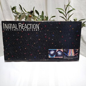 May include: A vintage board game box for "Initial Reaction: The Famous Name Game." The box is black with white text, featuring the game's title and a list of names. The box has colorful graphics on the side.