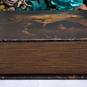 May include: A rectangular, antique-style box with a hinged lid. The lid is dark brown with a distressed, mottled pattern. The sides of the box have a wood-grain appearance. The box sits on a black base.