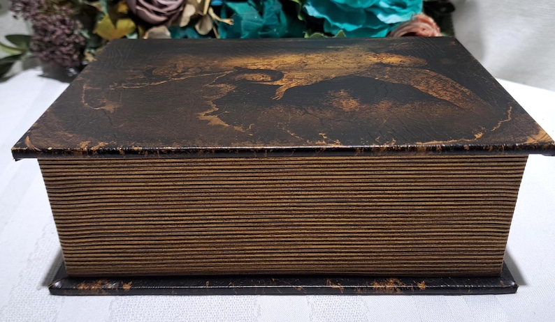 May include: A rectangular, antique-style box with a hinged lid. The lid is dark brown with a distressed, mottled pattern. The sides of the box have a wood-grain appearance. The box sits on a black base.