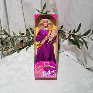 May include: A Barbie doll wearing a purple graduation gown and a gold sash that says "Class of 1997". She is holding a bouquet of flowers and wearing a graduation cap. The doll is in a pink and white box that says "Special Edition Graduation Barbie".
