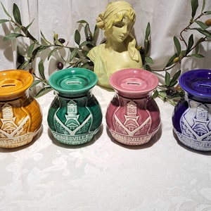 May include: Four ceramic piggy banks in different colors: brown, green, pink, and blue. Each bank features a white design of a building with a clock tower and the text "ZANESVILLE, OHIO".