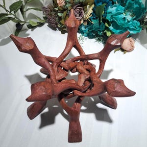 May include: A handcrafted, reddish-brown wooden decorative stand. The stand features an intricate, interwoven design with six stylized animal head shapes extending outwards. The base is a single, sturdy stem. The stand is likely used for displaying decorative items.