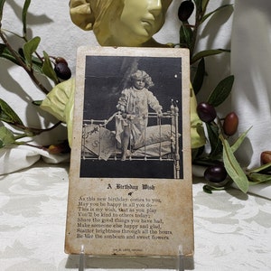 May include: A vintage postcard with a black and white photo of a young girl standing in a bed. The postcard has a birthday wish written on it that reads: "As this new birthday comes to you, May you be happy in all you do, This is my wish, that as you play You'll be kind to others today; Share the good things you have had, Make someone else happy and glad, Scatter brightness through all the hours Be like the sunbeam and sweet flowers."