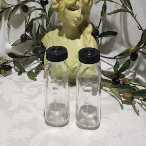 May include: Two clear glass baby bottles with black caps and markings. The bottles are on a white surface with a decorative bust in the background.