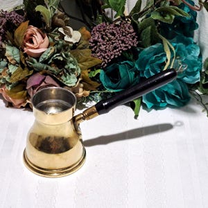 May include: A brass coffee pot with a black wooden handle. The pot has a rounded base and a cylindrical top. The handle is attached with a brass fitting. The pot is likely used for making Turkish or Greek coffee.