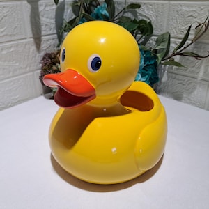 May include: A bright yellow duck-shaped container with a smooth, glossy finish. The duck features a vibrant orange beak and two dark blue eyes. The top of the duck's back has a wide, open circular cavity, making it functional as a storage holder.