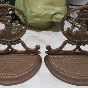 May include: A pair of antique brass bookends featuring an anchor design. The bookends have a rope-like border around the anchor and a decorative base.