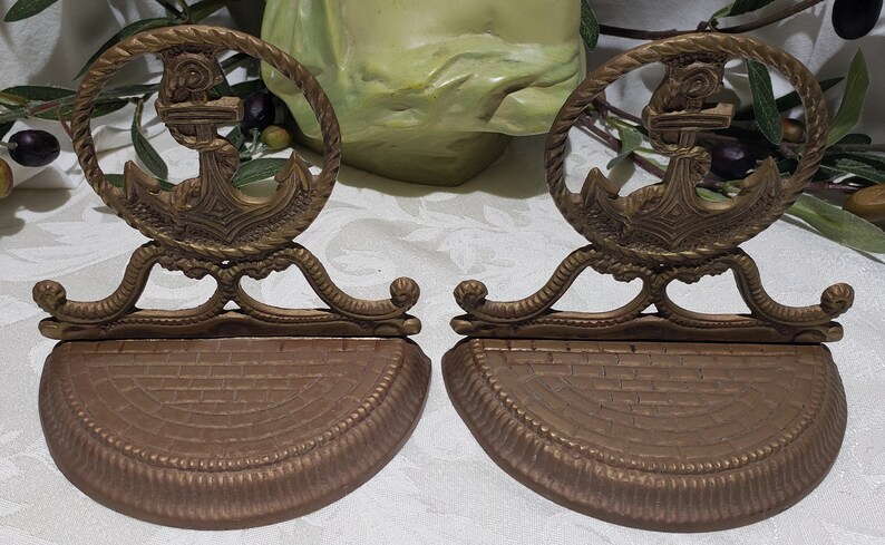 May include: A pair of antique brass bookends featuring an anchor design. The bookends have a rope-like border around the anchor and a decorative base.