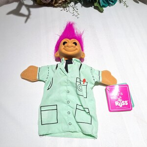 May include: A vintage Troll doll with bright pink hair and a smiling face. The doll wears a light green lab coat with black outlines and a pocket with a red pen. A pink tag with the text "Say it with a RUSS" is attached.
