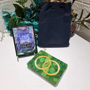 May include: A deck of tarot cards with a green marble-patterned back featuring two interlocking gold rings, accompanied by a single upright card titled Fairy Festival Imbolc and a dark blue leather drawstring pouch for storage.