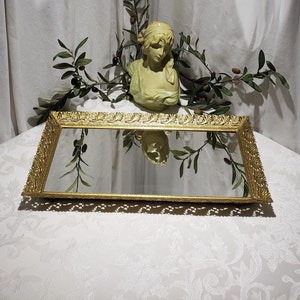 May include: A gold-toned decorative tray with a beveled mirror surface and an ornate, scrolled frame. The tray is sitting on a white surface.