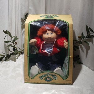 May include: A Cabbage Patch Kids doll in a red jacket, blue jeans, and white sneakers. The doll has red hair and blue eyes. The doll is in a box with the Cabbage Patch Kids logo and the text "I'm one of a kind. I'm Adoptable." and "Ages 3 & up".