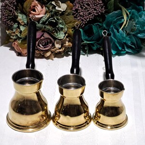 May include: Three brass coffee pots with black wooden handles. The pots have a classic design with a wide base and a narrow top. The handles have a loop at the end for hanging. The background features a floral arrangement.