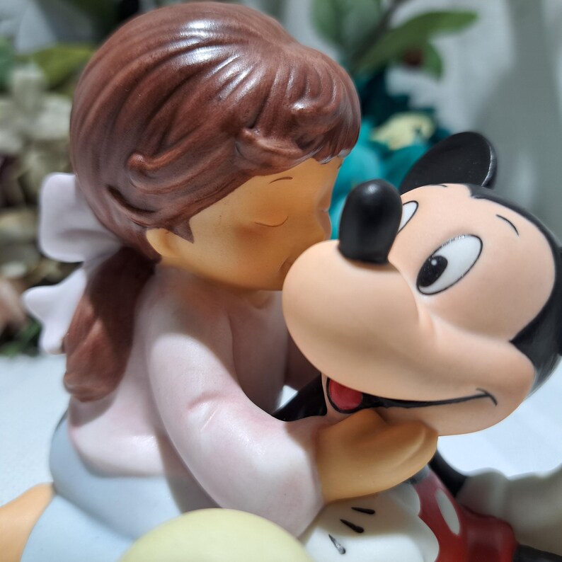 Pode incluir: Uma estatueta de cer&acirc;mica com uma menina de vestido rosa e la&ccedil;o branco beijando Mickey Mouse. A estatueta tem pele clara, cabelo castanho e olhos fechados. Mickey Mouse est&aacute; com sua roupa cl&aacute;ssica.
