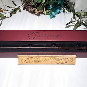 May include: A dark brown wand with a detailed handle rests in a burgundy box. The box is open, revealing a black interior. The box lid reads "Ollivanders" in gold lettering. A small rectangular sign below reads "Diagon Alley".