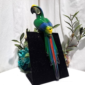 May include: A decorative parrot figurine featuring a vibrant green body, a blue and yellow head, and a long, multi-colored tail with red, blue, and green feathers. The bird is mounted on a black display stand, showcasing its detailed painted finish.