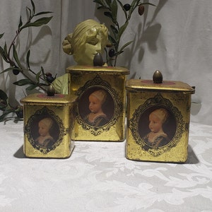 May include: Three gold-colored metal boxes with ornate designs and portraits of children. The boxes have hinged lids and are decorated with gold accents.