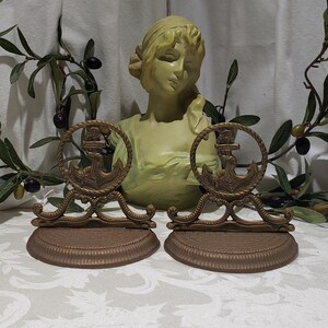 May include: A pair of gold-toned metal bookends featuring an anchor design. The bookends are on a white surface with a green bust in the background.