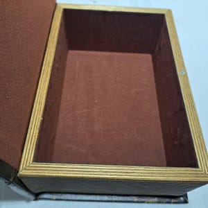 May include: Open, rectangular box with a brown interior and a gold-colored, textured frame. The box is lined with a dark brown fabric. The exterior appears to be a dark brown, possibly leather-like material. The box is open, revealing the empty interior.