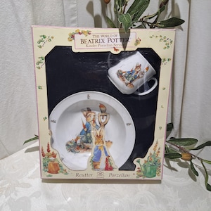 May include: A boxed set of Beatrix Potter children's dinnerware. The set includes a bowl, a plate, and a mug. Each piece features a colorful illustration of Peter Rabbit and Benjamin Bunny. The box is white with green and yellow accents and the text "The World of Beatrix Potter Kinder Porzellan Service" and "Reutter Porzellan".