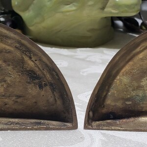 May include: A pair of brass bookends in the shape of a half-circle. The bookends are worn and have a patina finish. The bottom of one bookend has a sticker that reads "ANTIQUE FINISHED  MADE IN INDIA  C.O. 3104".