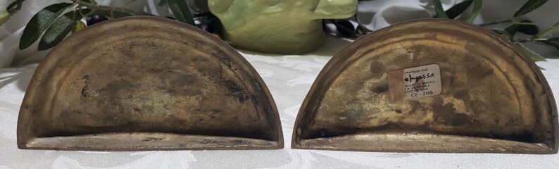 May include: A pair of brass bookends in the shape of a half-circle. The bookends are worn and have a patina finish. The bottom of one bookend has a sticker that reads "ANTIQUE FINISHED  MADE IN INDIA  C.O. 3104".