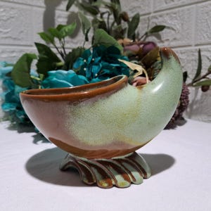 May include: A ceramic planter with a horn-shaped silhouette, featuring a two-tone glaze of light mint green and warm brown. The vessel rests on a wide, scalloped base with radiating ridges, showcasing a smooth, glossy finish throughout the piece.