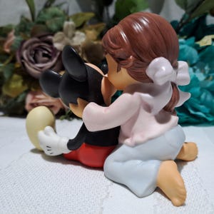 Pode incluir: Uma estatueta retratando Mickey Mouse sendo abra&ccedil;ado por uma menina. Mickey est&aacute; com sua roupa cl&aacute;ssica, e a menina usa um top rosa, cal&ccedil;as azul claro e um la&ccedil;o branco. A estatueta &eacute; colocada em um fundo de flores.