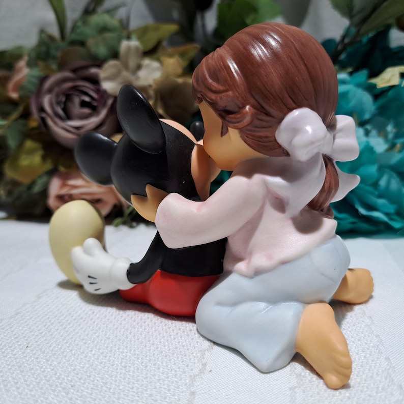 Pode incluir: Uma estatueta retratando Mickey Mouse sendo abra&ccedil;ado por uma menina. Mickey est&aacute; com sua roupa cl&aacute;ssica, e a menina usa um top rosa, cal&ccedil;as azul claro e um la&ccedil;o branco. A estatueta &eacute; colocada em um fundo de flores.