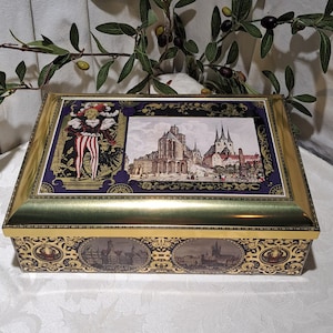 May include: A gold and purple rectangular tin box with a gold trim. The lid features a picture of a building with two towers and a man in a colorful costume. The bottom of the box has a repeating pattern of buildings.