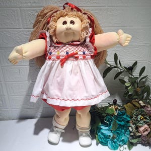 May include: A soft-sculpture doll with light brown yarn hair in pigtails, wearing a white dress with red smocking, red floral embroidery, and a red ribbon tie. The doll has light skin, white shoes, and red bloomers, with arms extended outward.